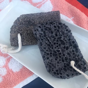 May include: Two black pumice stones with white string handles on a white rectangular plate. The plate is on top of a pink and white towel.