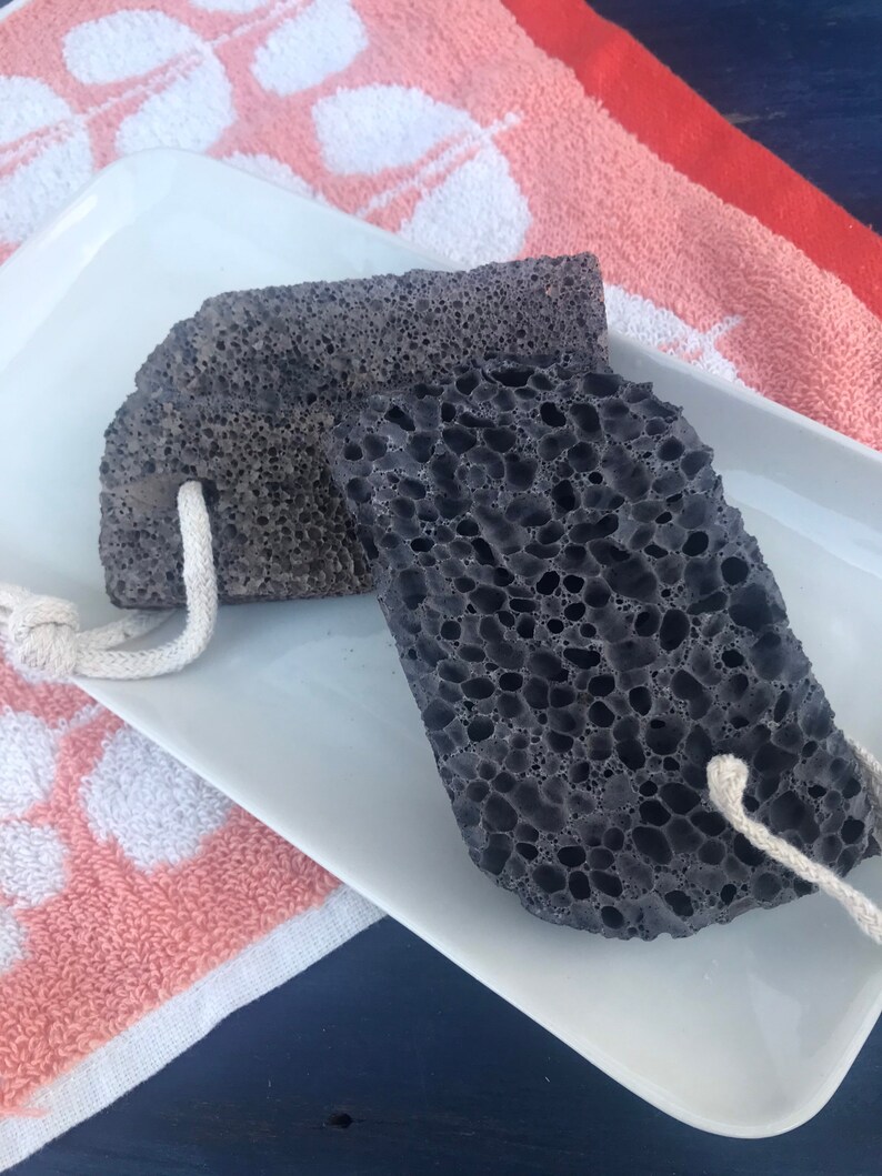 May include: Two black pumice stones with white string handles on a white rectangular plate. The plate is on top of a pink and white towel.