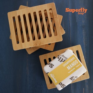 May include: Three wooden soap dishes stacked on top of each other. The top dish has a bar of mango shampoo soap on it. The soap is wrapped in white paper with black text that reads "Superfly Soap Shampoo Bar Mango handmade - vegan".