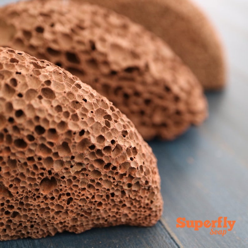May include: Three brown pumice stones with a rough, porous texture. The stones are arranged in a row on a blue surface. The text "Superfly Soap" is visible in the lower right corner of the image.