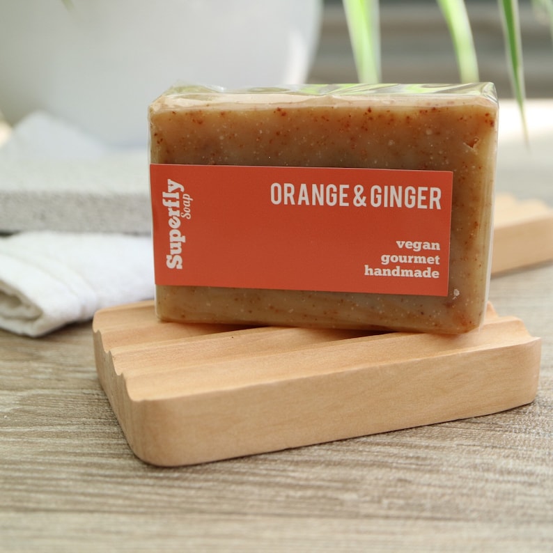 Puede incluir: Una pastilla de jab&oacute;n de naranja y jengibre en un envoltorio de pl&aacute;stico transparente. El jab&oacute;n est&aacute; sobre un jabonera de madera. El jab&oacute;n est&aacute; etiquetado como "Superfly Soap" y "Naranja & Jengibre". La etiqueta tambi&eacute;n dice "vegano, gourmet, hecho a mano".
