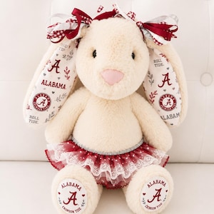 May include: A cream-colored plush rabbit toy featuring long ears with Alabama Crimson Tide branding, including the letter A, the text Roll Tide, and floral accents. The rabbit wears a layered red and white lace tutu and a decorative bow.