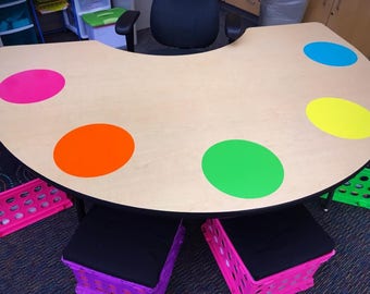 May include: A round wooden table with colorful circles on the surface. The table is surrounded by four pink and black storage bins.