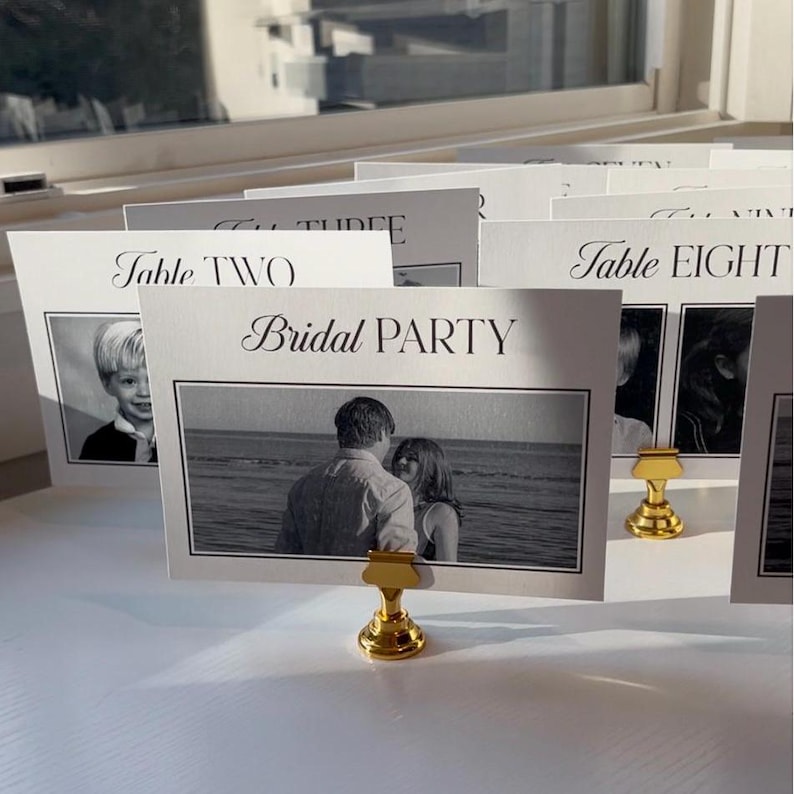 May include: Black and white wedding table number cards with gold holders. The cards feature photos and the text "Table Two", "Bridal Party", and "Table Eight".
