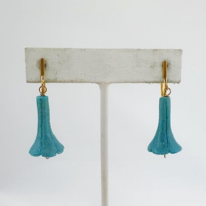 May include: A pair of turquoise blue flower-shaped earrings with gold-colored ear wires. The earrings are displayed on a white earring stand against a white background. The earrings have a textured surface and a delicate, floral design.