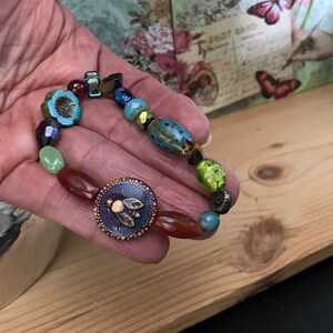 May include: A beaded bracelet featuring a variety of colorful beads, including turquoise flower-shaped beads, faceted dark purple beads, and a large blue button with a bee design. The bracelet is held in a hand.