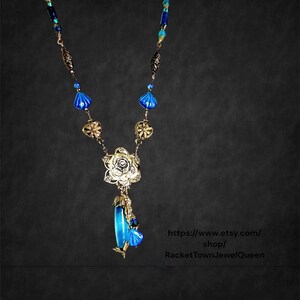 May include: A blue and gold necklace with a floral pendant and a long, teardrop-shaped blue bead. The necklace is made of metal and glass beads. The pendant features a rose design. The necklace is available for purchase on Etsy from the shop RacketTownJewelQueen.