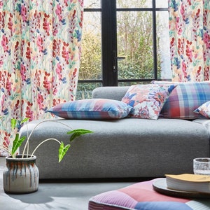 Peut inclure: Un canapé gris avec des coussins décoratifs à motifs colorés, une plante en pot et un livre sur un petit pouf. Le canapé est devant une fenêtre avec des rideaux à motifs floraux.