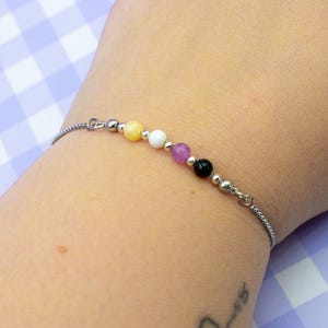May include: A silver chain bracelet with six colorful beads: yellow, white, purple, black, and two silver beads. The bracelet is on a wrist against a purple and white checkered background.