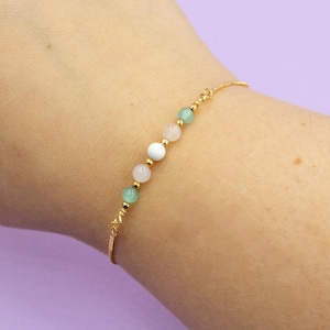 May include: A delicate bracelet with a gold chain and five round beads in shades of green, white, and pink.