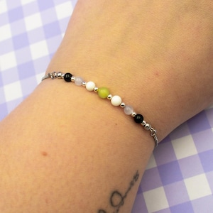 May include: A silver chain bracelet with a series of colorful beads. The beads include black, white, light green, and pink. The bracelet is displayed on a wrist against a purple and white checkered background.