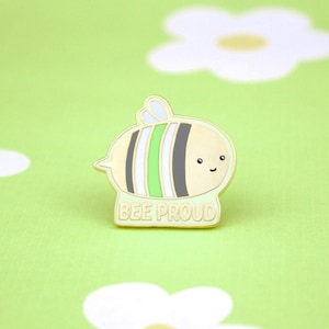 May include: A gold-colored enamel pin featuring a cartoon bee with green, white, and black stripes. The bee has a smiling face and the text "BEE PROUD" is written below it.