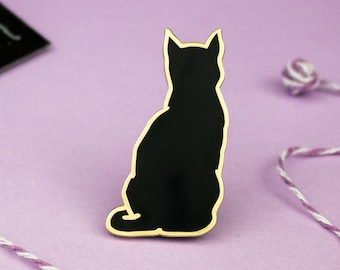 Op de afbeelding: Zwarte kat silhouet emaille pin met een gouden rand. De pin is op een paarse achtergrond met wit en paars touw.