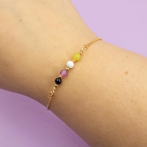 Könnte beinhalten: Ein goldenes Kettenarmband mit vier Perlen in den Farben der Non-Binary-Pride-Flagge: Gelb, Weiß, Lila und Schwarz.