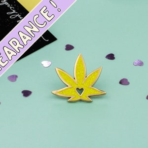 May include: A gold-coloured pin with a yellow glitter marijuana leaf design. The leaf has a heart cut out of the centre.