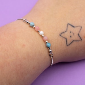 May include: A delicate bracelet with pink, white, and blue beads, interspersed with small silver beads, on a silver chain. The background is a solid purple. The bracelet is a piece of jewellery.