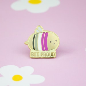 Könnte beinhalten: Eine goldfarbene Emaille-Anstecknadel mit einer Cartoon-Biene mit einem geschlechtsneutralen Pride-Flaggen-Design. Die Biene hat ein fröhliches Gesicht und der Text "BEE PROUD" ist darunter geschrieben.
