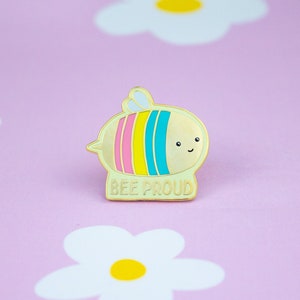 May include: A gold-coloured enamel pin featuring a cartoon bee with a pink, yellow and blue striped body. The bee has a smiling face and the text "BEE PROUD" is written below it.