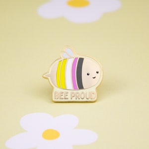 Puede incluir: Pin de abeja dorada con rayas amarillas, blancas, rosas y negras. La abeja tiene una cara sonriente y el texto "BEE PROUD" está debajo.