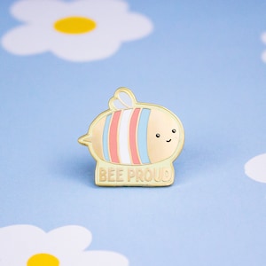 以下が含まれることがあります： トランスジェンダーのプライドフラッグ柄の漫画の蜂が描かれた金色のエナメルピン。蜂は笑顔で、「BEE PROUD」という文字が下に書かれています。