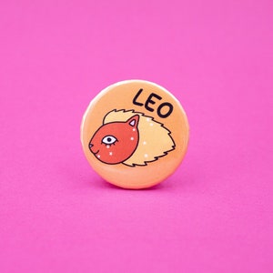 Puede incluir: Un botón redondo naranja con la cabeza de un león de dibujos animados y la palabra "LEO" impresa en él.