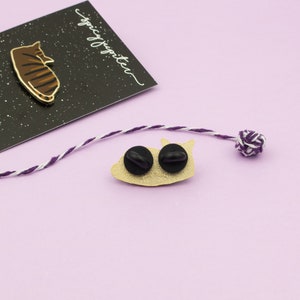 May include: Gold-colored enamel pin of a cat curled up sleeping. The pin has a black starry background and the text "spicy kitten" is written on the back. The pin has two black rubber stoppers on the back.