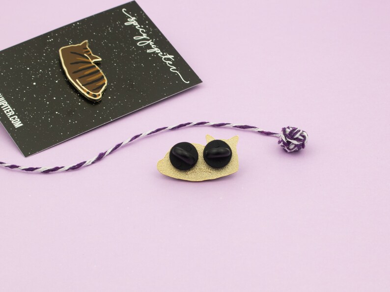 May include: Gold-colored enamel pin of a cat curled up sleeping. The pin has a black starry background and the text "spicy kitten" is written on the back. The pin has two black rubber stoppers on the back.