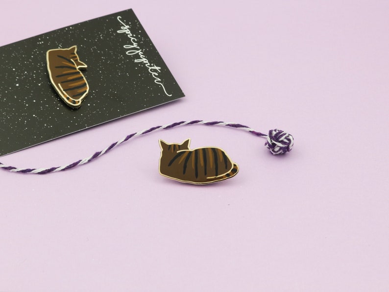 May include: Two gold enamel pins featuring a brown tabby cat in different poses. The pins are on a purple background with a white and purple string and a small ball of string.