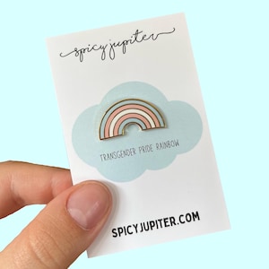 Op de afbeelding: Transgender Pride Rainbow emaille pin op een witte kaart met de tekst "spicy jupiter" en "SPICYJUPITER.COM". De pin is een regenboog met roze, witte en blauwe strepen.