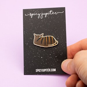 May include: A brown and black enamel pin of a cat laying down on a black background with white speckles. The pin has a white outline and the text "spicyjupiter.com" is printed at the bottom of the card.