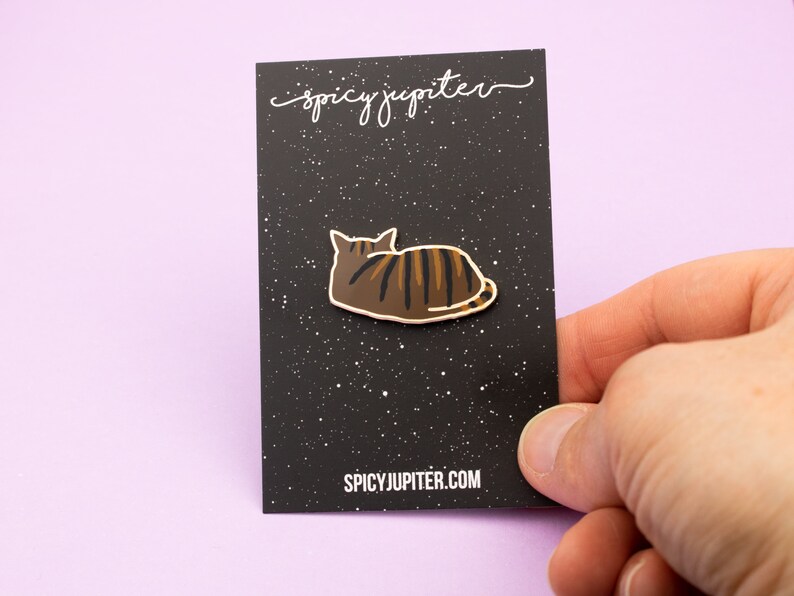 May include: A brown and black enamel pin of a cat laying down on a black background with white speckles. The pin has a white outline and the text "spicyjupiter.com" is printed at the bottom of the card.
