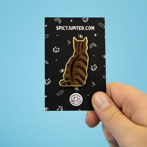May include: A gold-colored enamel pin featuring a brown tabby cat sitting with its back to the viewer. The pin is on a black card with white text that reads "SPICYJUPITER.COM".