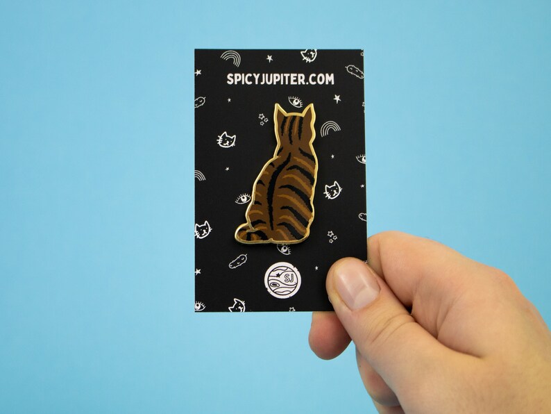 May include: A gold-colored enamel pin featuring a brown tabby cat sitting with its back to the viewer. The pin is on a black card with white text that reads "SPICYJUPITER.COM".