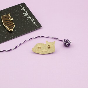 May include: Gold-colored enamel pin of a cat curled up sleeping. The back of the pin is shown, with two metal posts for attaching to fabric. The pin is on a light purple surface.