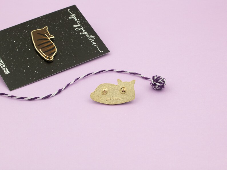 May include: Gold-colored enamel pin of a cat curled up sleeping. The back of the pin is shown, with two metal posts for attaching to fabric. The pin is on a light purple surface.