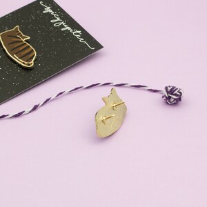 May include: A gold-colored enamel pin featuring a cartoon cat lying down. The pin is on a black background with white stars. The text "spicy jupiter" is written in white on the black background. The back of the pin is shown with a gold finish and a safety pin closure.