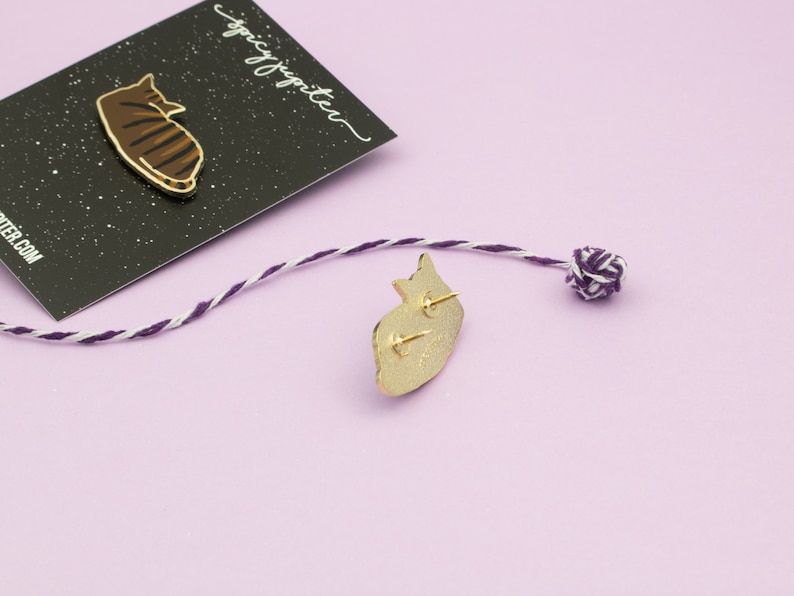 May include: A gold-colored enamel pin featuring a cartoon cat lying down. The pin is on a black background with white stars. The text "spicy jupiter" is written in white on the black background. The back of the pin is shown with a gold finish and a safety pin closure.