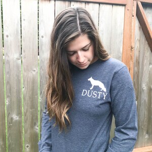 K&ouml;nnte beinhalten: Eine Frau tr&auml;gt ein dunkelblaues Langarmshirt mit einer wei&szlig;en Silhouette eines Hundes und dem Wort "DUSTY" auf der Vorderseite.