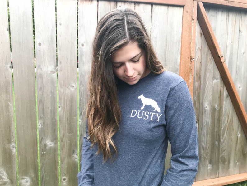 K&ouml;nnte beinhalten: Eine Frau tr&auml;gt ein dunkelblaues Langarmshirt mit einer wei&szlig;en Silhouette eines Hundes und dem Wort "DUSTY" auf der Vorderseite.