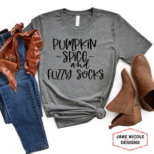 May include: Gray t-shirt with the text "Pumpkin Spice and Fuzzy Socks" in black lettering. The shirt is displayed on a white background with a pair of brown ankle boots and a brown and orange scarf.