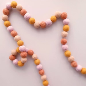 Pode incluir: Uma guirlanda de bolas de feltro em tons de rosa, laranja e amarelo. A guirlanda é enfiada com um fio branco e é perfeita para adicionar um toque de cor a qualquer ambiente.