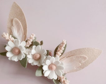 May include: A felt flower crown with white daisies, green leaves, and pink felt accents. The crown is decorated with two beige felt bunny ears.