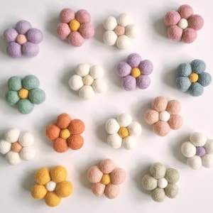 May include: A selection of colourful felt flower decorations in shades of pink, purple, green, orange, and yellow. Each flower features rounded petals around a central sphere. The flowers are arranged on a white surface.