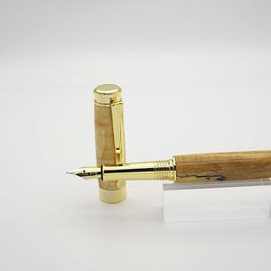 May include: A fountain pen with a gold-colored nib and accents. The pen's barrel is made of light-colored wood with dark graining. The cap is also light-colored wood with a gold clip. The pen is displayed on a clear acrylic stand.
