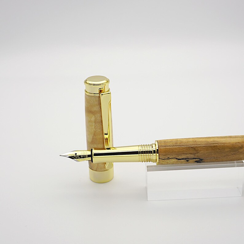May include: A fountain pen with a gold-colored nib and accents. The pen's barrel is made of light-colored wood with dark graining. The cap is also light-colored wood with a gold clip. The pen is displayed on a clear acrylic stand.