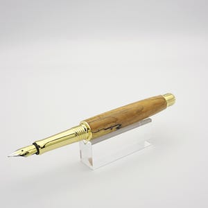 May include: A handcrafted fountain pen with a light brown wooden barrel and gold-colored accents. The pen features a gold nib and is displayed on a clear acrylic stand. The pen's design showcases a blend of natural wood grain and metallic elements.
