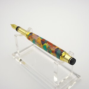 May include: A gold and black fountain pen with a colorful, multi-colored, abstract resin body. The pen is on a clear acrylic stand.