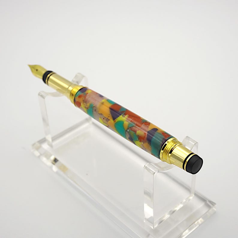 May include: A gold and black fountain pen with a colorful, multi-colored, abstract resin body. The pen is on a clear acrylic stand.