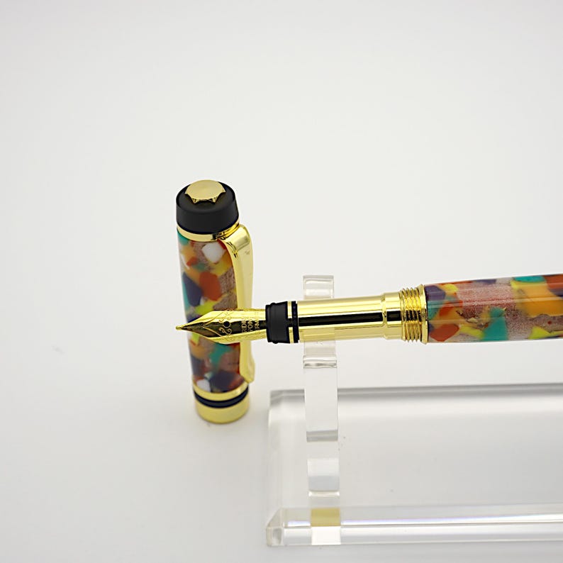 May include: A gold and black fountain pen with a colorful, marbled resin body. The pen is on a clear acrylic stand.