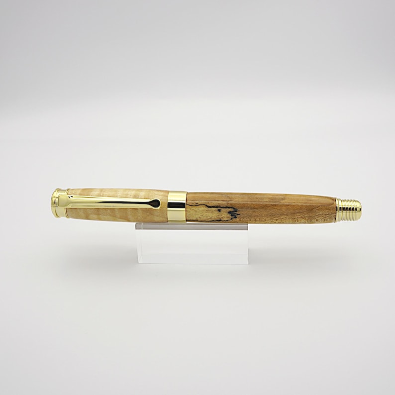 May include: A handcrafted wooden pen with a light-colored, patterned barrel and a darker, solid wood section. The pen features gold-colored metal accents and a gold-colored nib. The pen is displayed on a clear acrylic stand against a white background.